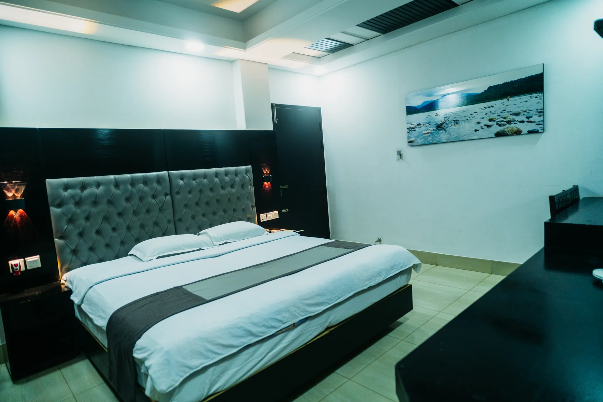 Sky Lawn Hotel's guest bedroom offers a modern, spacious design with a large tufted headboard bed...